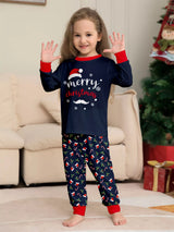 Girl wearing matching navy Merry Christmas pajama sets with red cuffs and holiday-print pants beside a decorated Christmas tree.