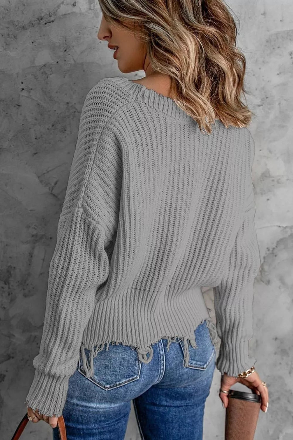Woman wearing a gray ribbed V-neck sweater with distressed raw hem and blue jeans.