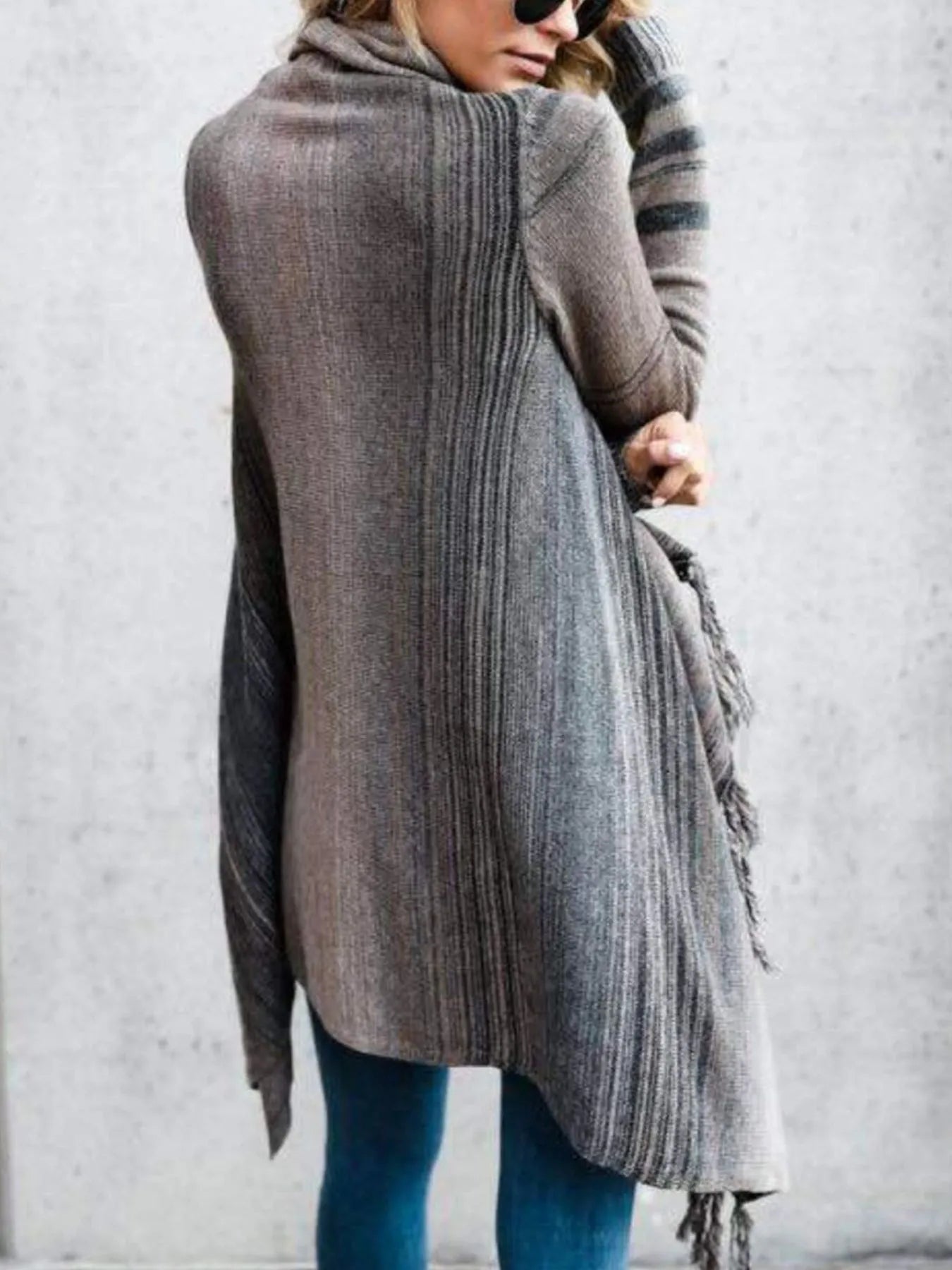 Woman wearing a gray stripe fringe long sleeve wrap cardigan with an asymmetrical draped front over a white top and jeans.