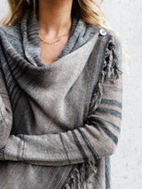 Woman wearing a gray stripe fringe long sleeve wrap cardigan with an asymmetrical draped front over a white top and jeans.