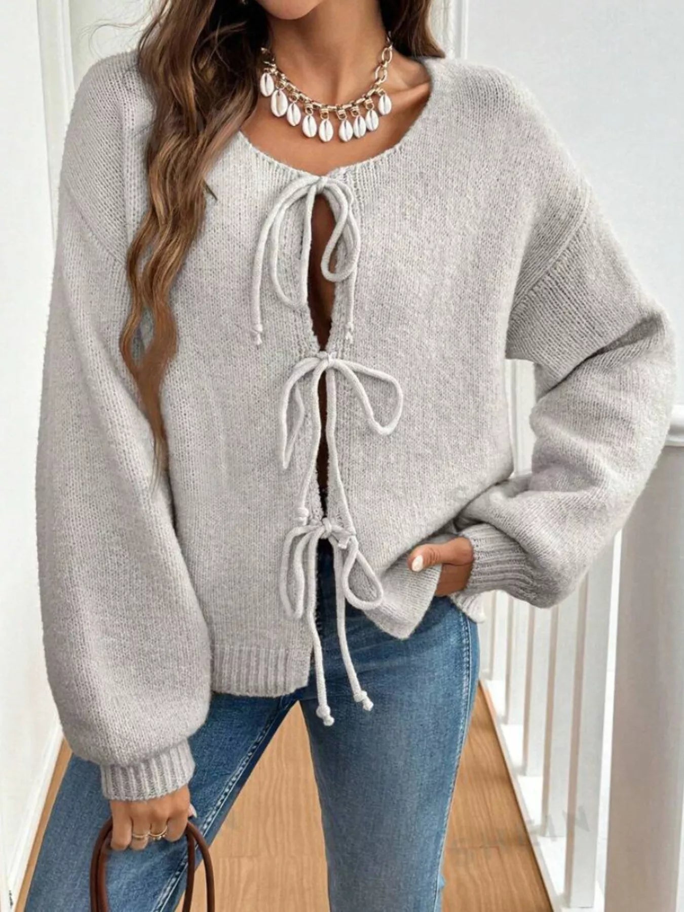 Woman wearing a gray tie-front long sleeve knit cardigan with three bow ties paired with blue jeans.