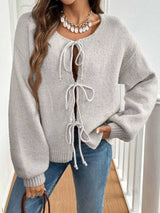 Woman wearing a gray tie-front long sleeve knit cardigan with three bow ties paired with blue jeans.