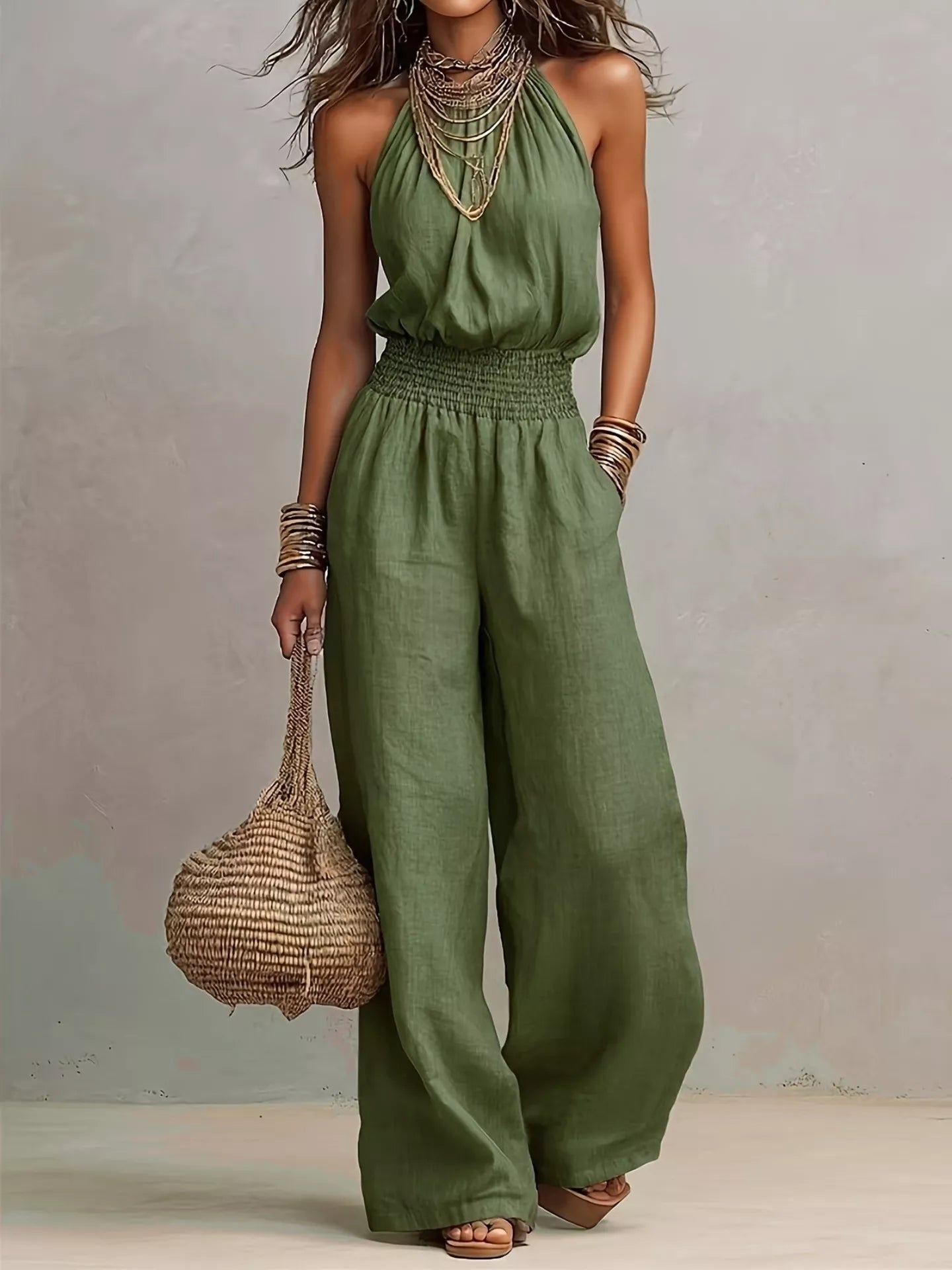 Woman wearing a green halter-neck wide-leg jumpsuit with a smocked waist, layered gold jewelry, and bracelets, styled for an elegant evening look.