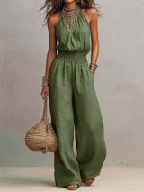 Woman wearing a green halter-neck wide-leg jumpsuit with a smocked waist, layered gold jewelry, and bracelets, styled for an elegant evening look.