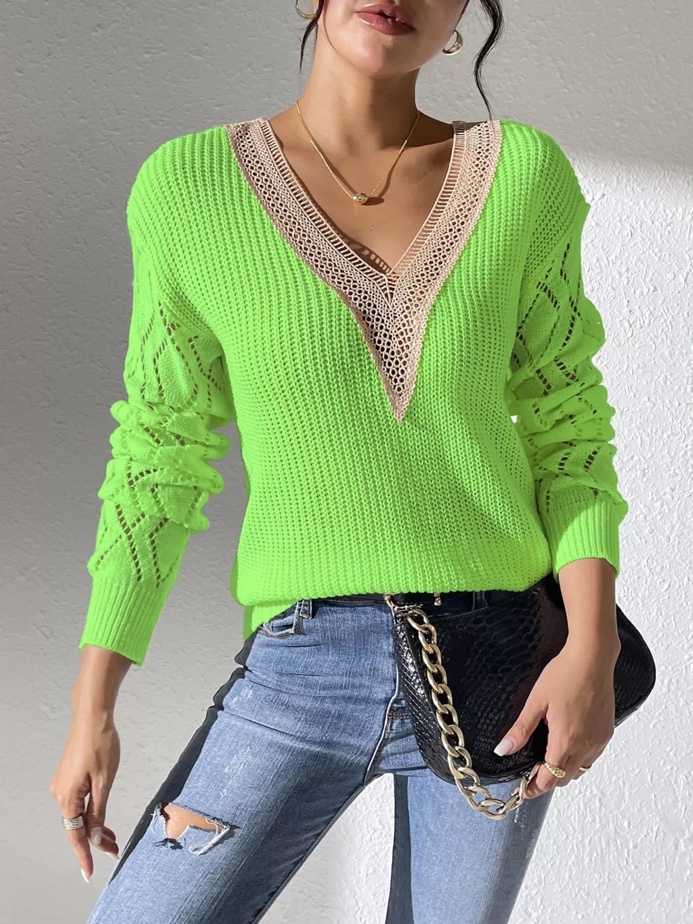 Woman wearing a green V-neck hollow knit sweater with contrasting neckline, openwork long sleeves, and blue jeans.