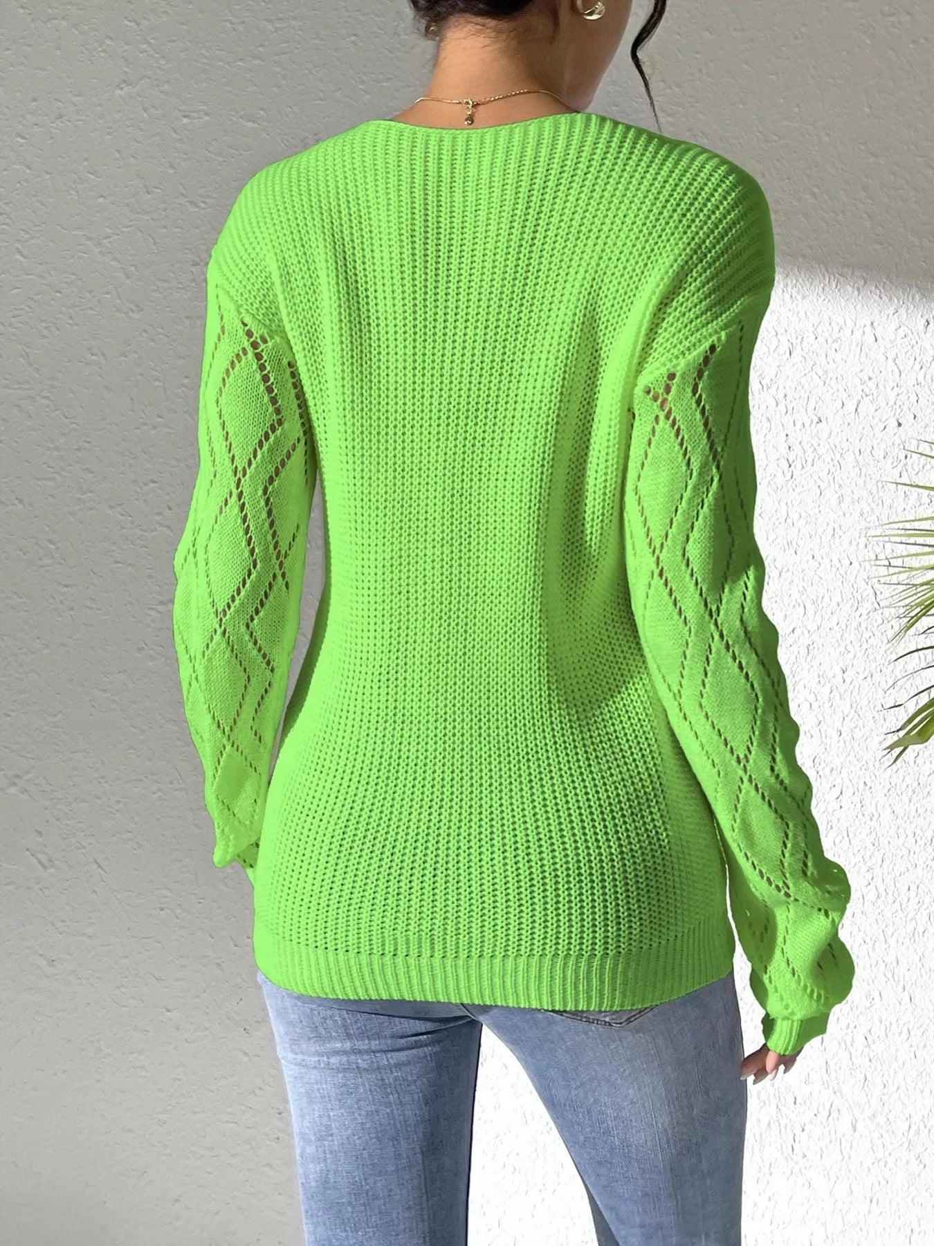 Woman wearing a green V-neck hollow knit sweater with contrasting neckline, openwork long sleeves, and blue jeans.