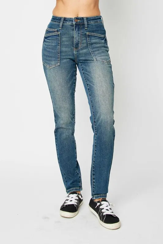 Model wearing Judy Blue high-waist skinny jeans in a medium wash with oversized front pockets and a slim, ankle-length fit, paired with black sneakers.