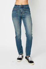 Model wearing Judy Blue high-waist skinny jeans in a medium wash with oversized front pockets and a slim, ankle-length fit, paired with black sneakers.