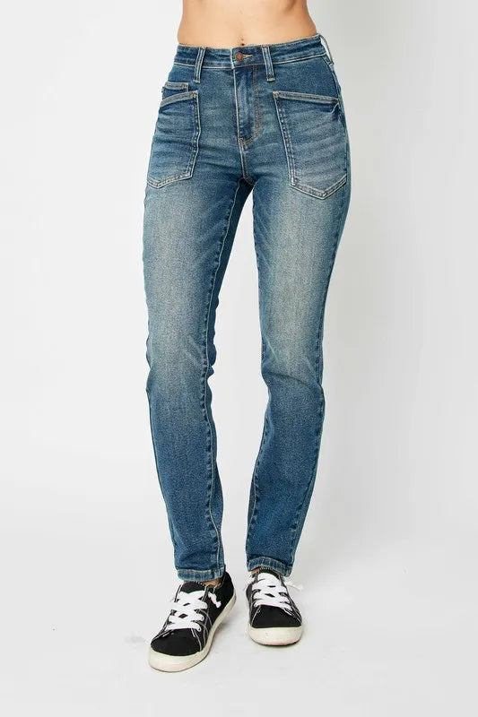 Model wearing Judy Blue high-waist skinny jeans in a medium wash with oversized front pockets and a slim, ankle-length fit, paired with black sneakers.