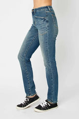 Model wearing Judy Blue high-waist skinny jeans in a medium wash with oversized front pockets and a slim, ankle-length fit, paired with black sneakers.
