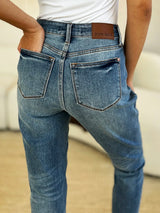 Judy Blue Full Size Mid Rise Rigid Magic Release Hem Jeans in medium wash featuring a flattering mid-rise fit, straight-leg silhouette, moderate stretch, and trendy raw release hem, styled with white sneakers.