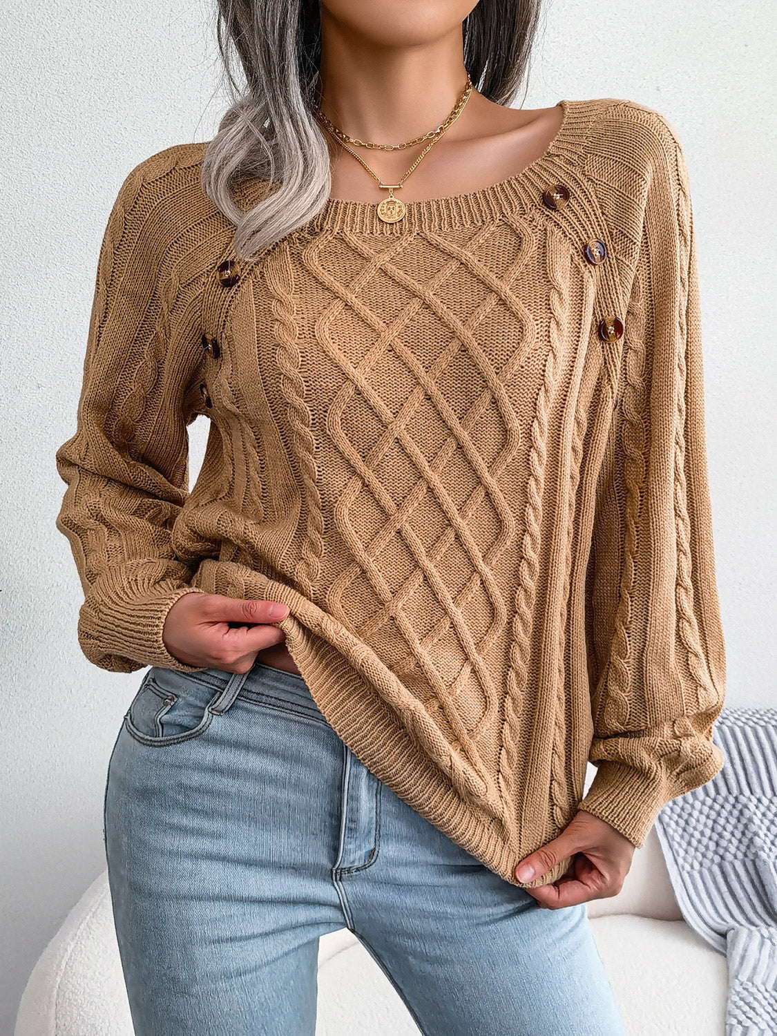 Woman wearing an khaki cable-knit sweater with decorative buttons, paired with light-wash jeans, layered gold necklaces, and a white handbag. Perfect for cozy fall and winter outfits.