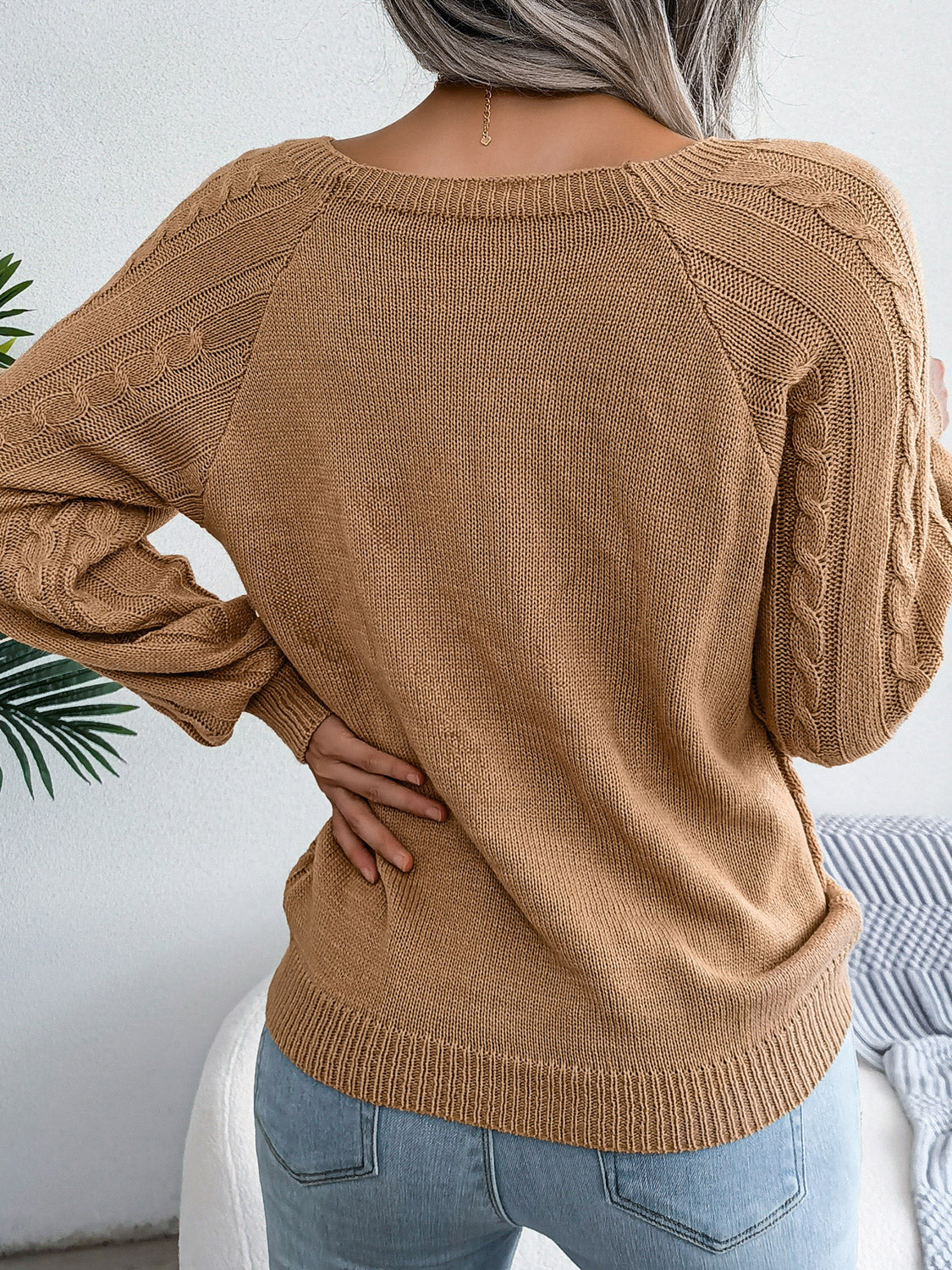 Woman wearing an khaki cable-knit sweater with decorative buttons, paired with light-wash jeans, layered gold necklaces, and a white handbag. Perfect for cozy fall and winter outfits.