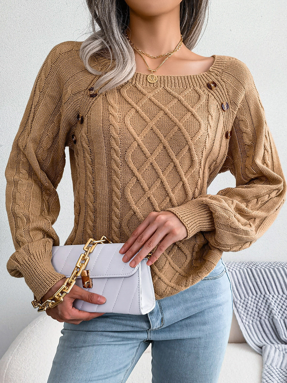 Woman wearing an khaki cable-knit sweater with decorative buttons, paired with light-wash jeans, layered gold necklaces, and a white handbag. Perfect for cozy fall and winter outfits.