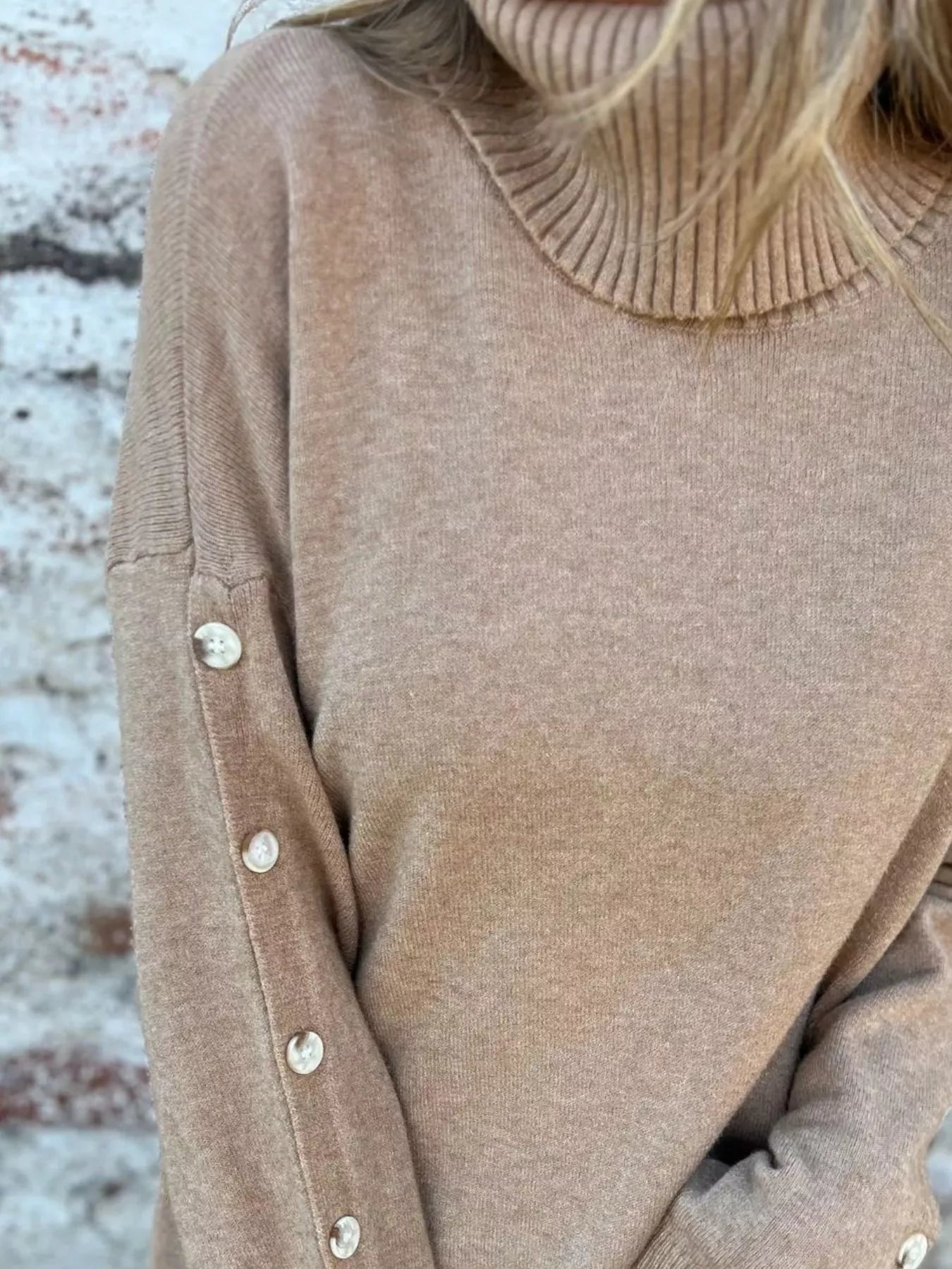Woman wearing a khaki ribbed turtleneck sweater with decorative button sleeves and jeans.