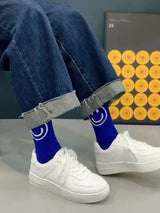 Person wearing blue socks with smiley face design, white sneakers, and rolled-up jeans against a gray wall with a decorative poster.
