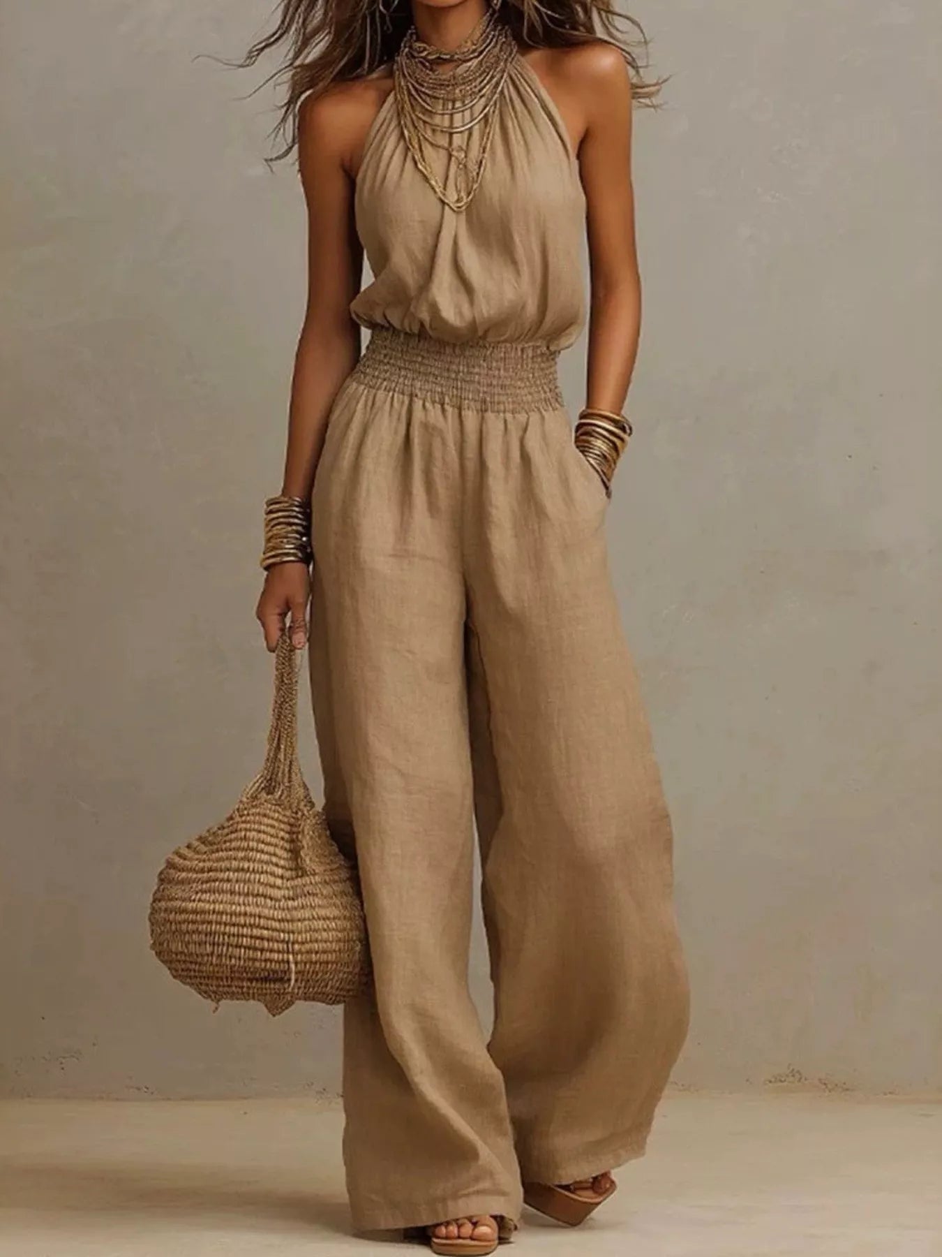 Woman wearing a light brown halter-neck wide-leg jumpsuit with a smocked waist, layered gold jewelry, and bracelets, styled for an elegant evening look.