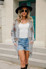 Woman wearing a light gray open-front cardigan with colorful star print, front pockets, denim shorts and black Western boots.