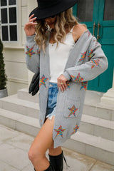 Woman wearing a light gray open-front cardigan with colorful star print, front pockets, denim shorts and black Western boots.