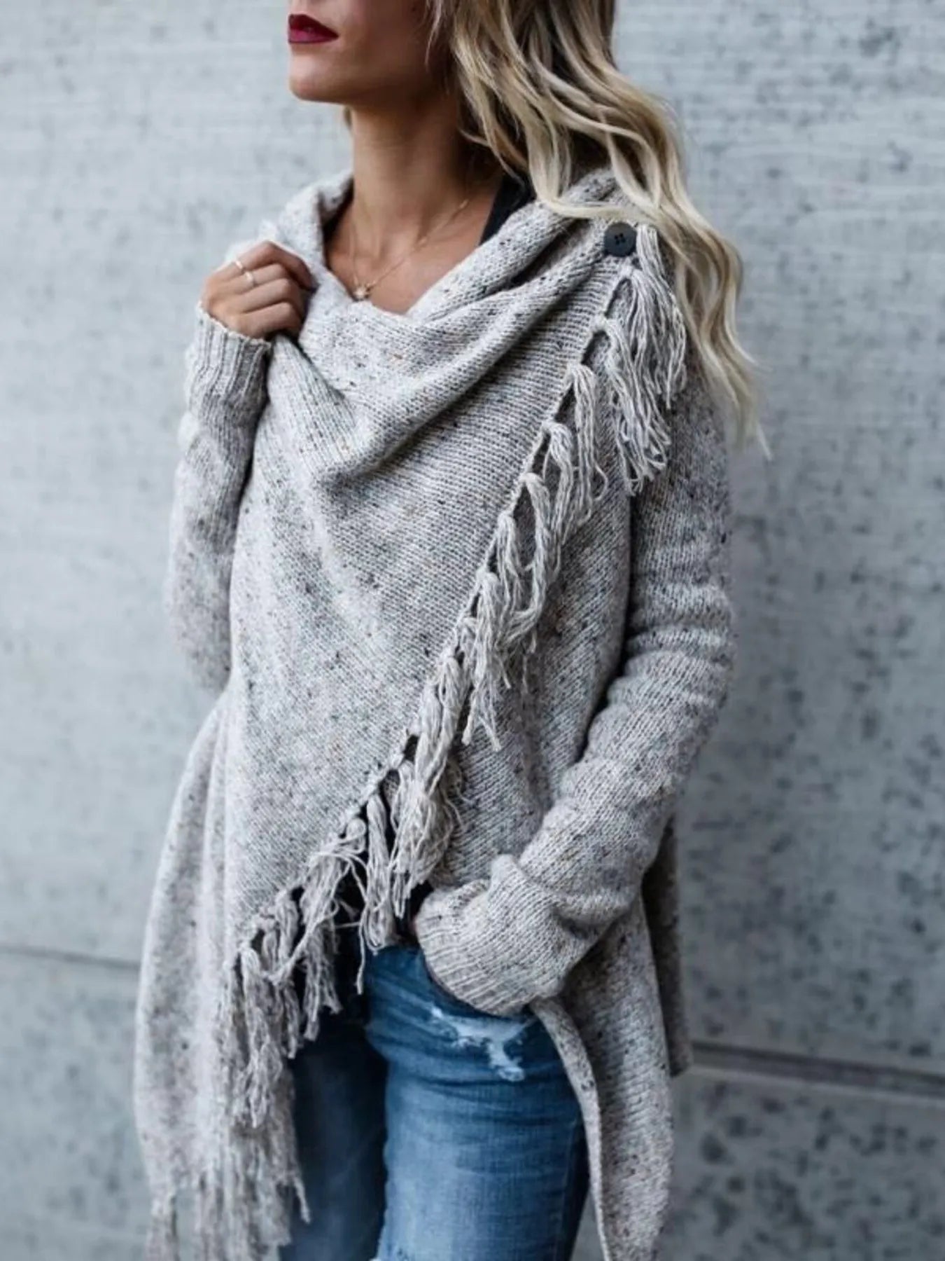 Woman wearing a light gray fringe long sleeve wrap cardigan with an asymmetrical draped front over a white top and jeans.