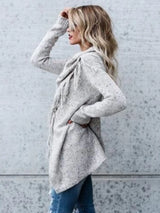 Woman wearing a light gray fringe long sleeve wrap cardigan with an asymmetrical draped front over a white top and jeans.
