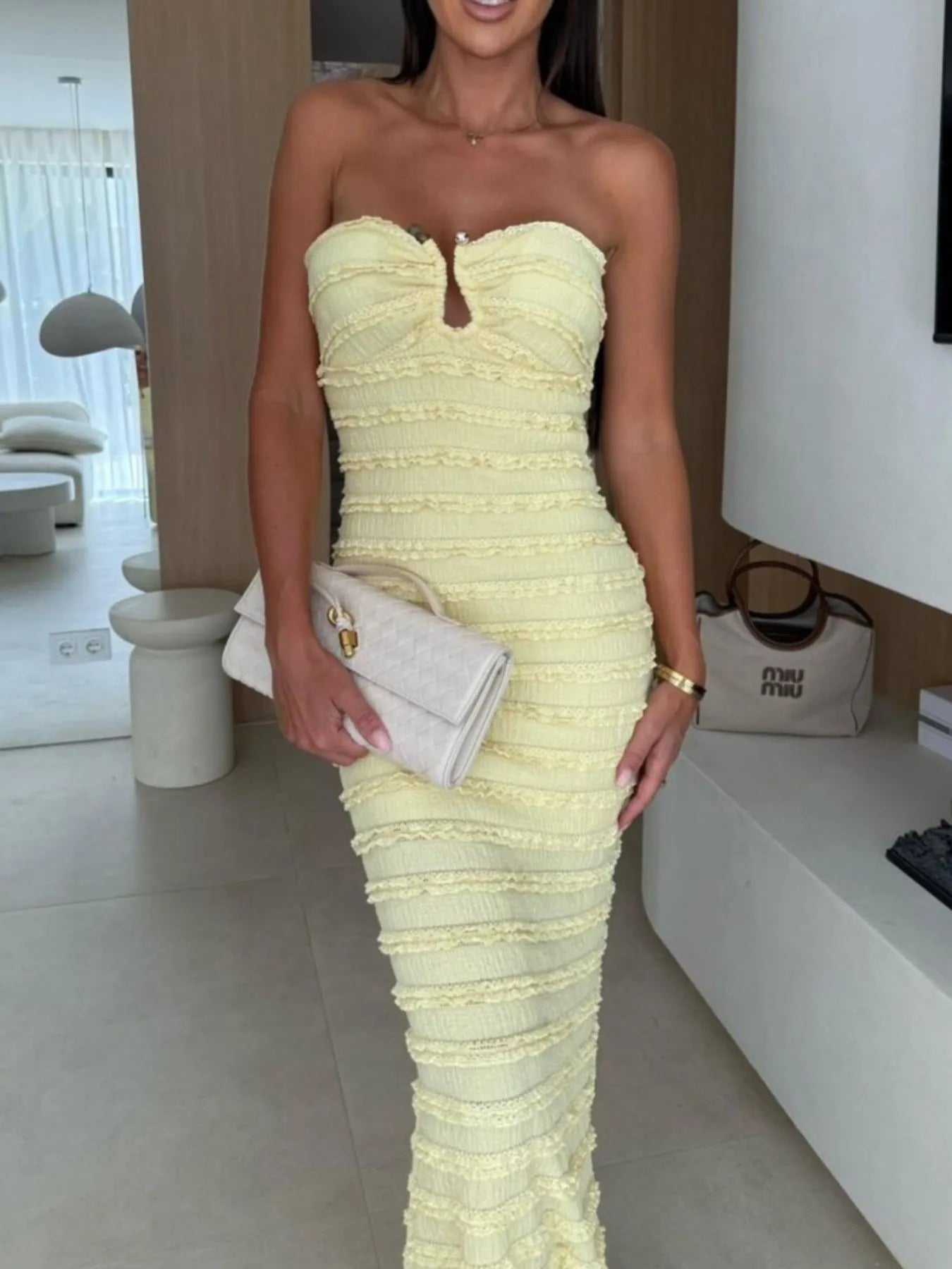 Woman wearing a light-yellow sweetheart neck tube maxi dress with ruched detailing and a subtle cutout, styled with an ivory clutch for an elegant evening look.