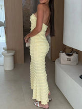 Woman wearing a light-yellow sweetheart neck tube maxi dress with ruched detailing and a subtle cutout, styled with an ivory clutch for an elegant evening look.
