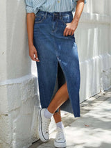 Woman wearing a medium wash denim midi skirt with front slit, pockets and raw hem.