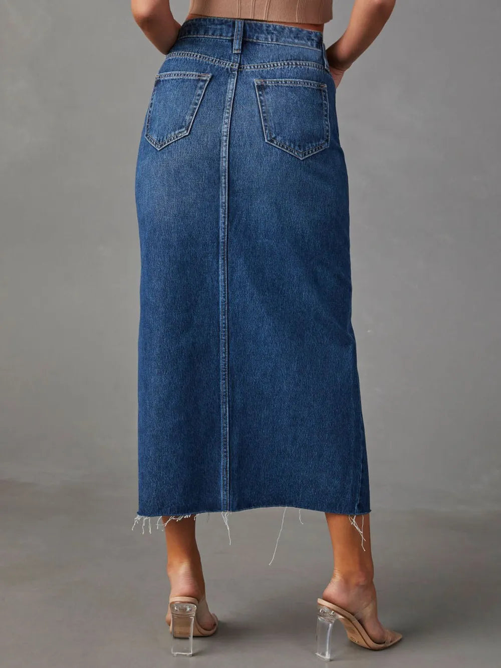 Woman wearing a medium wash denim midi skirt with front slit, pockets and raw hem.