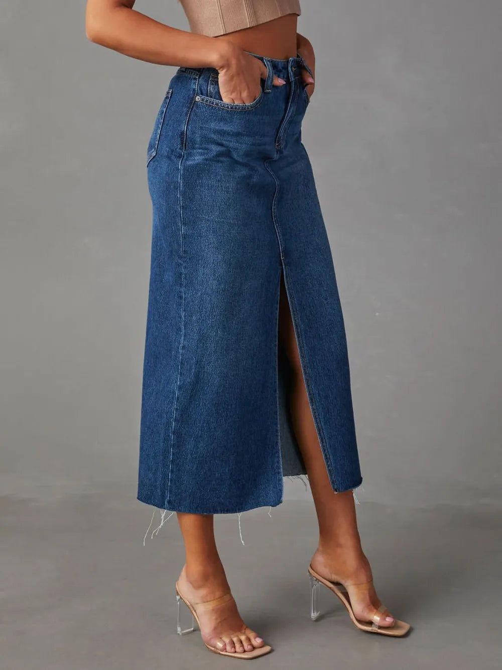 Woman wearing a medium wash denim midi skirt with front slit, pockets and raw hem.