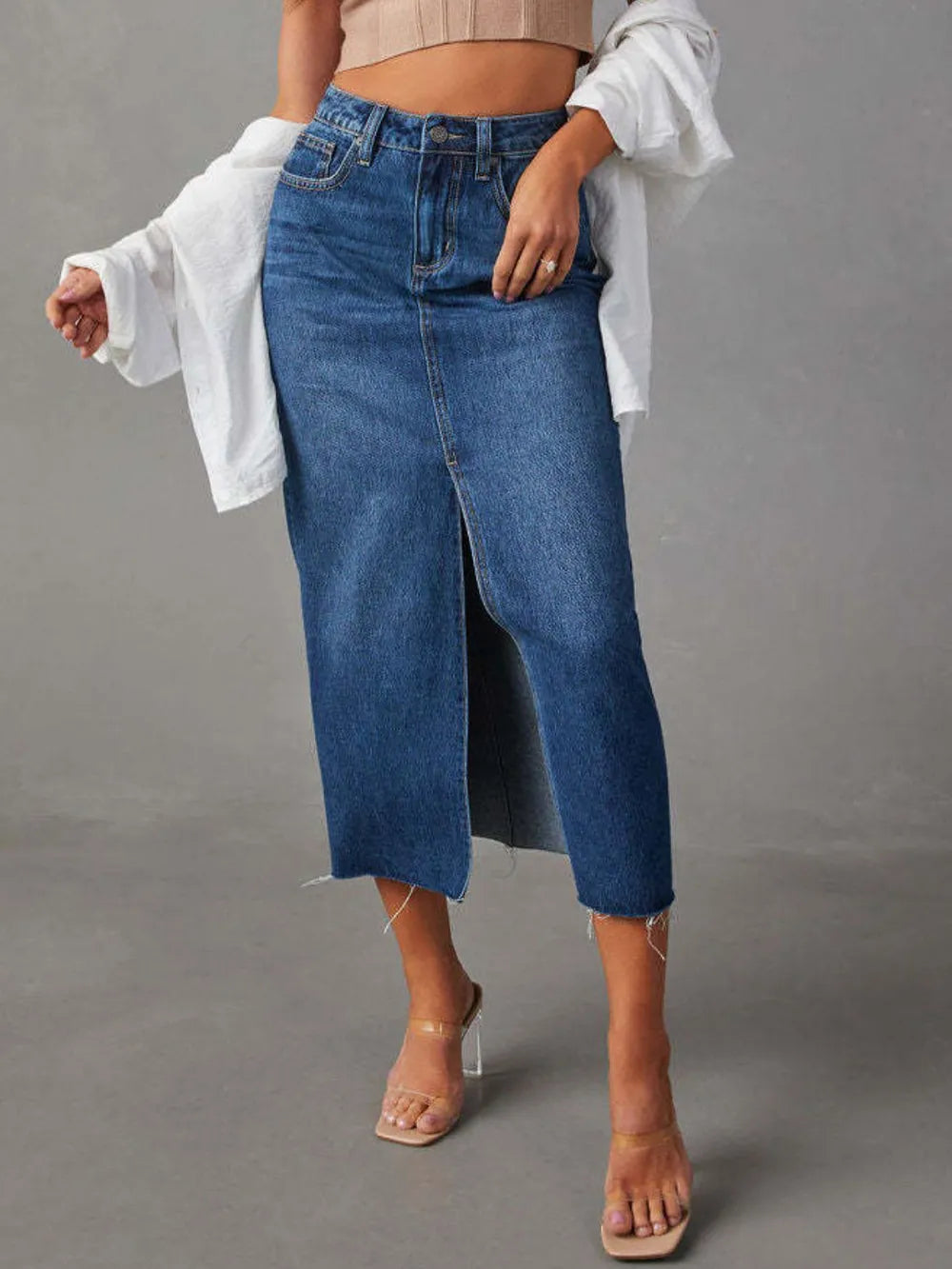Woman wearing a medium wash denim midi skirt with front slit, pockets and raw hem.
