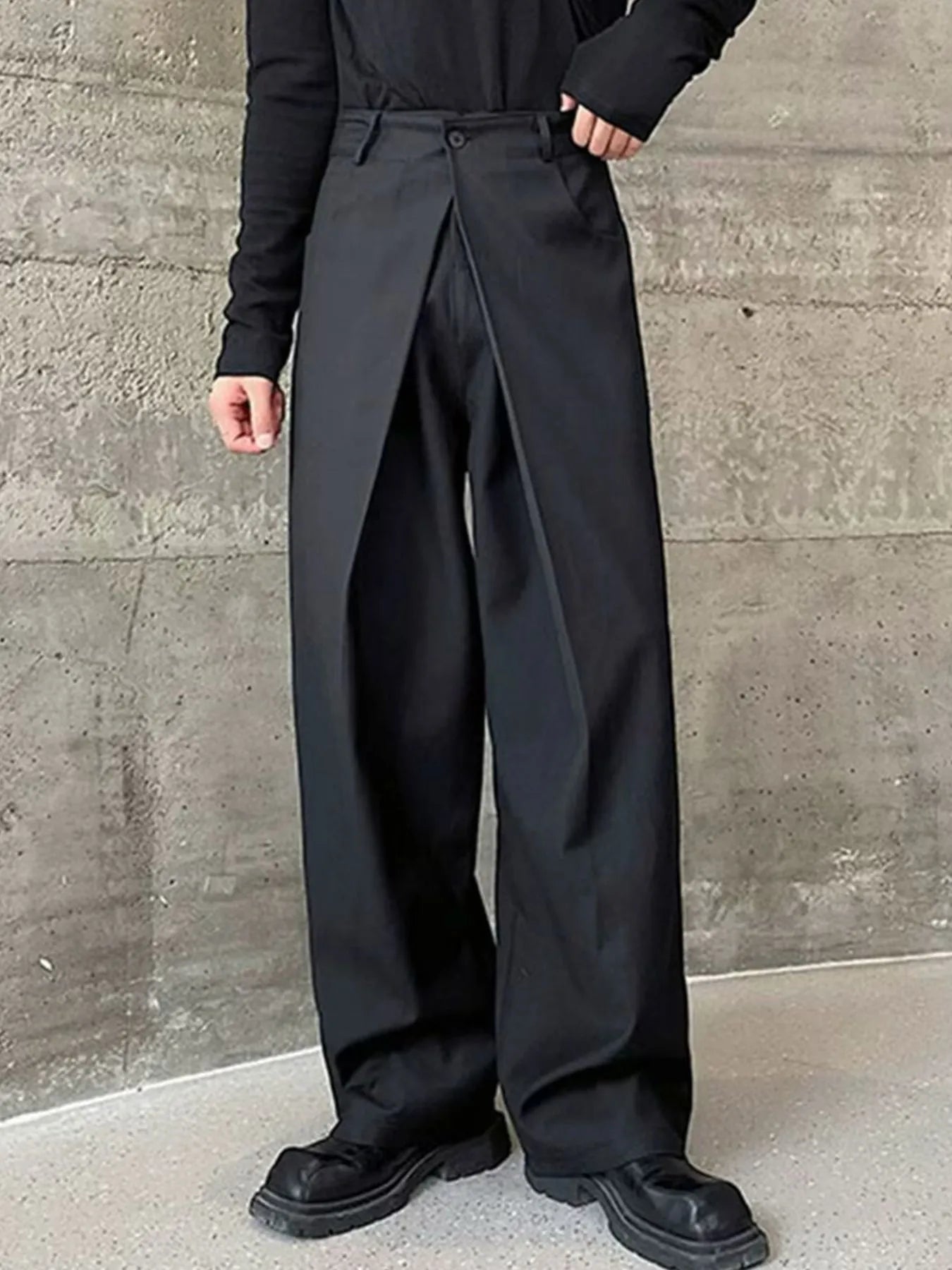 Men's black wide leg pleated pants with a button-front waistband and relaxed, full-length silhouette, styled with a black top and chunky black shoes.