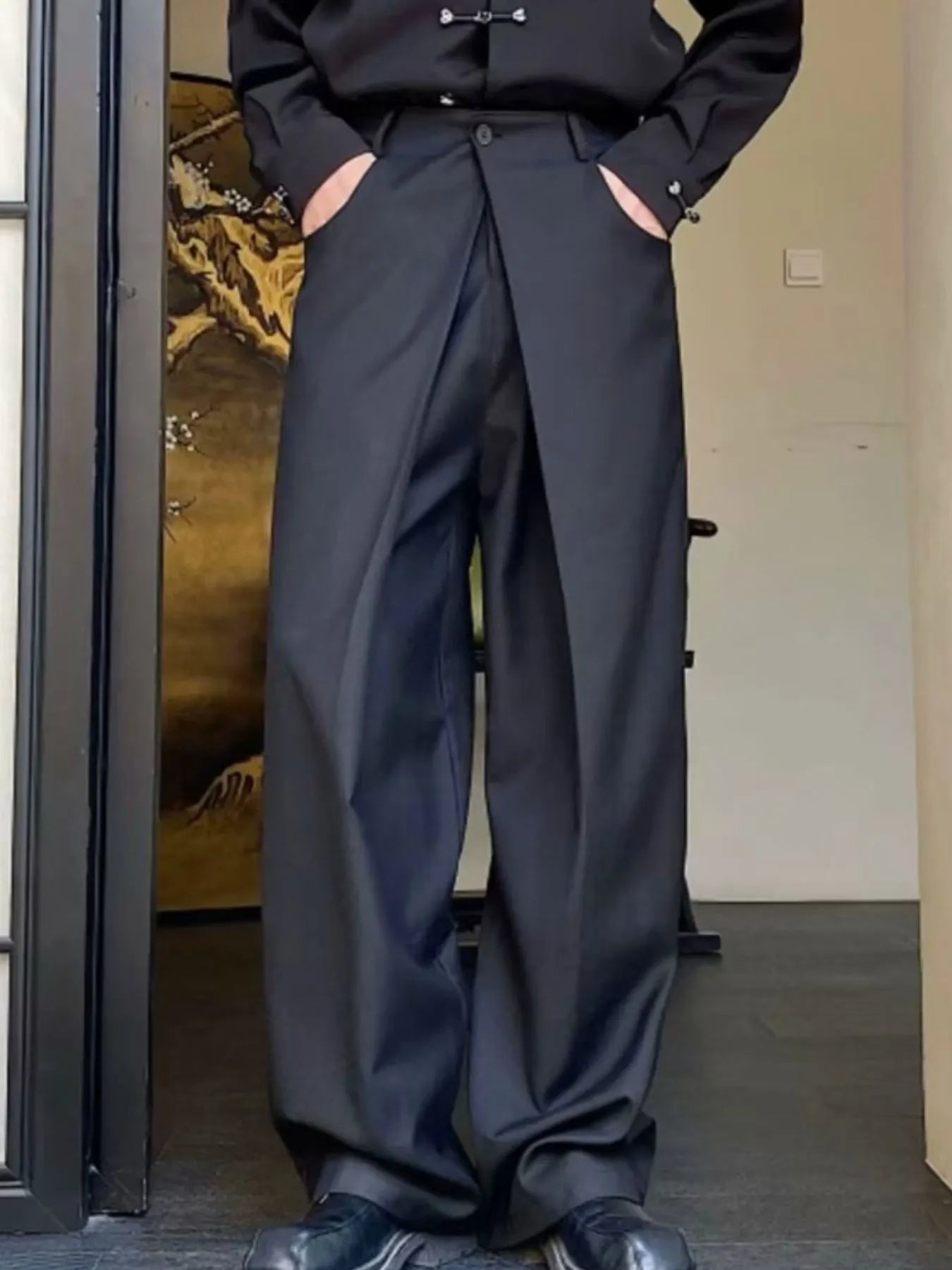 Men's black wide leg pleated pants with a button-front waistband and relaxed, full-length silhouette, styled with a black top and chunky black shoes.