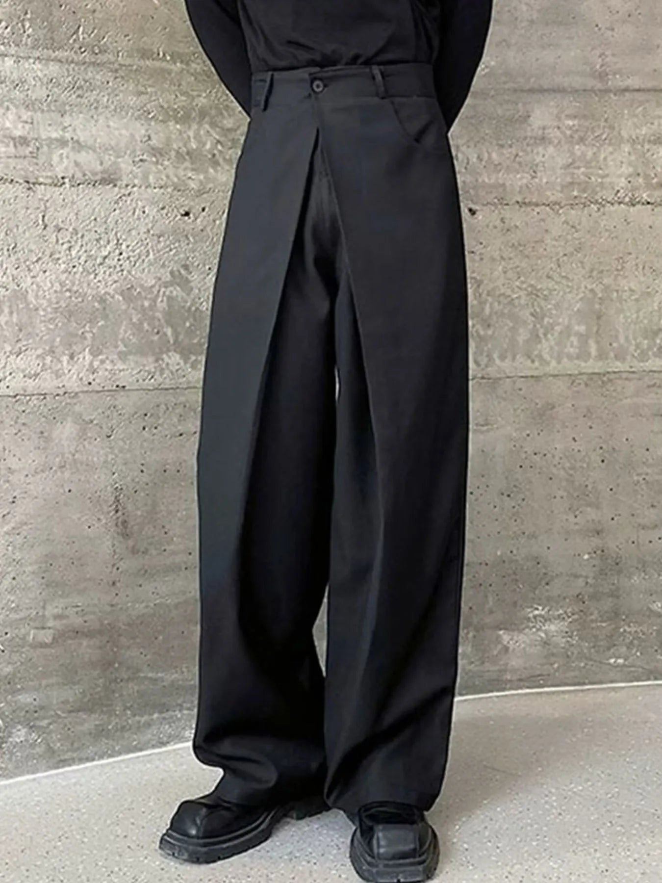 Men's black wide leg pleated pants with a button-front waistband and relaxed, full-length silhouette, styled with a black top and chunky black shoes.