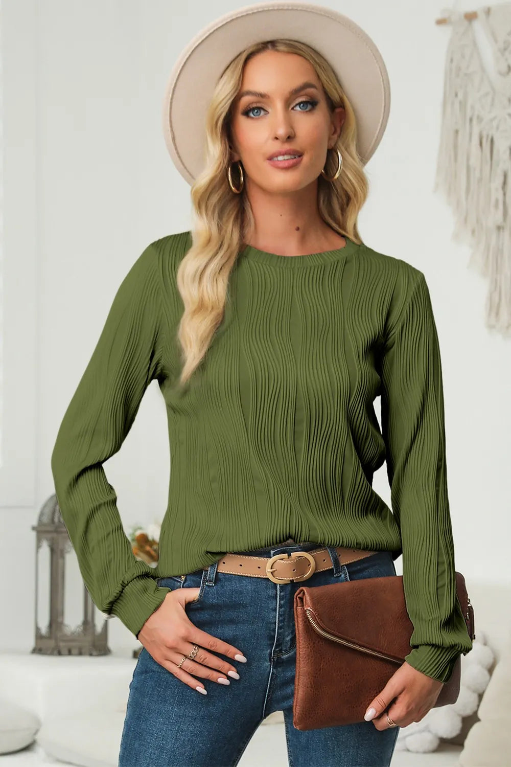 Woman wearing a moss textured round neck long sleeve blouse with dark-wash jeans, brown clutch and cream hat.