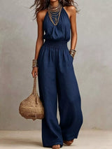 Woman wearing a navy blue halter-neck wide-leg jumpsuit with a smocked waist, layered gold jewelry, and bracelets, styled for an elegant evening look.