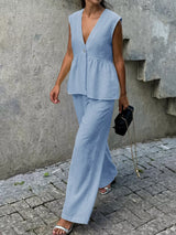 Woman wearing a light blue sleeveless top and pants against a concrete wall.