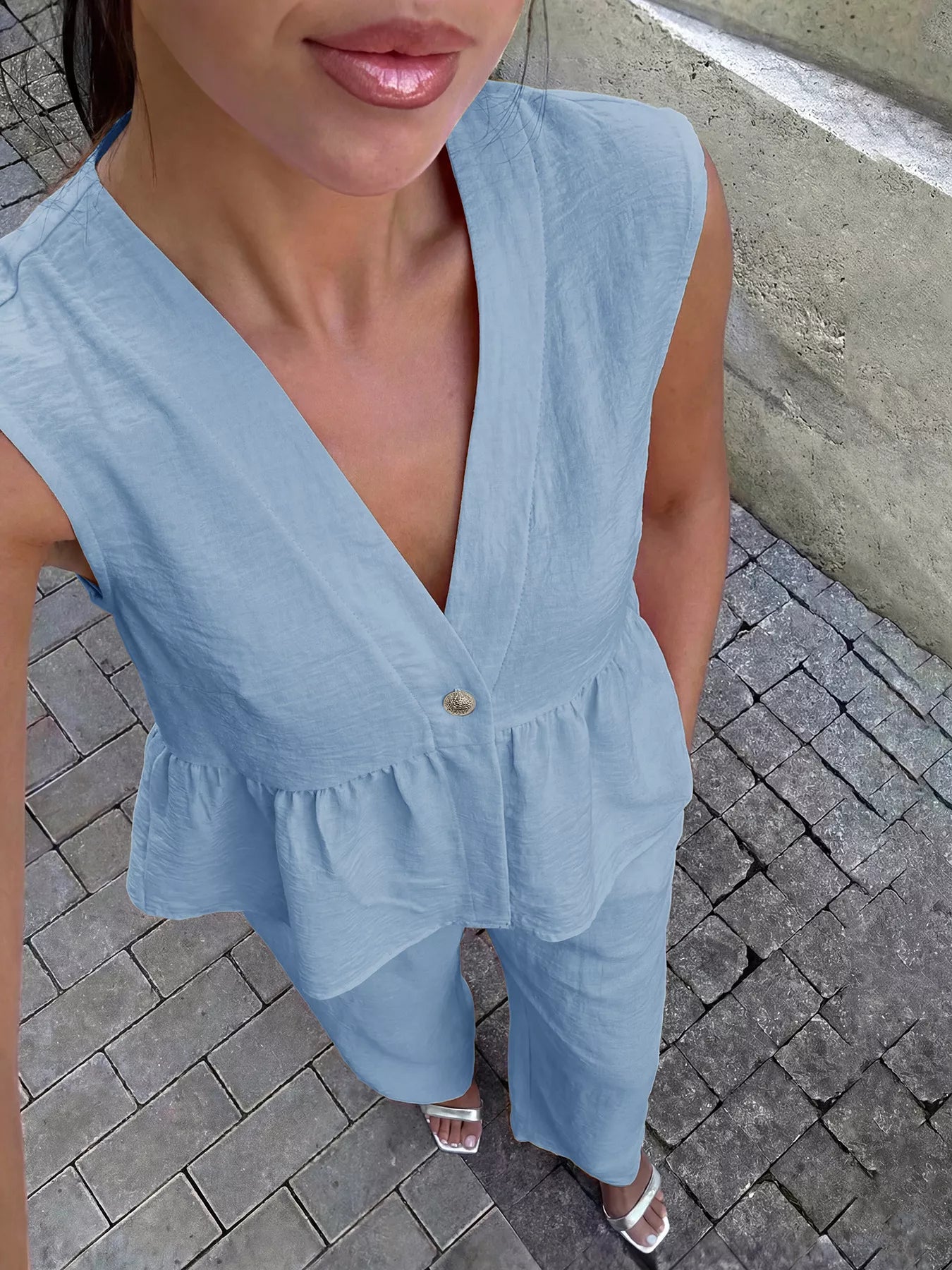 Woman wearing a light blue sleeveless top with a V-neck and ruffled details on a paved ground.