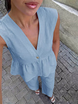 Woman wearing a light blue sleeveless top with a V-neck and ruffled details on a paved ground.