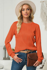 Woman wearing an orange textured round neck long sleeve blouse with dark-wash jeans, brown clutch and cream hat.