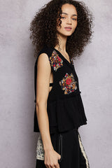 Woman wearing a black sleeveless top with floral embroidery against a gray background