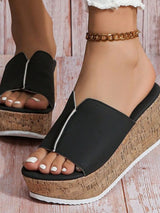 Black wedge sandals with cork soles worn by a person on a wooden floor.
