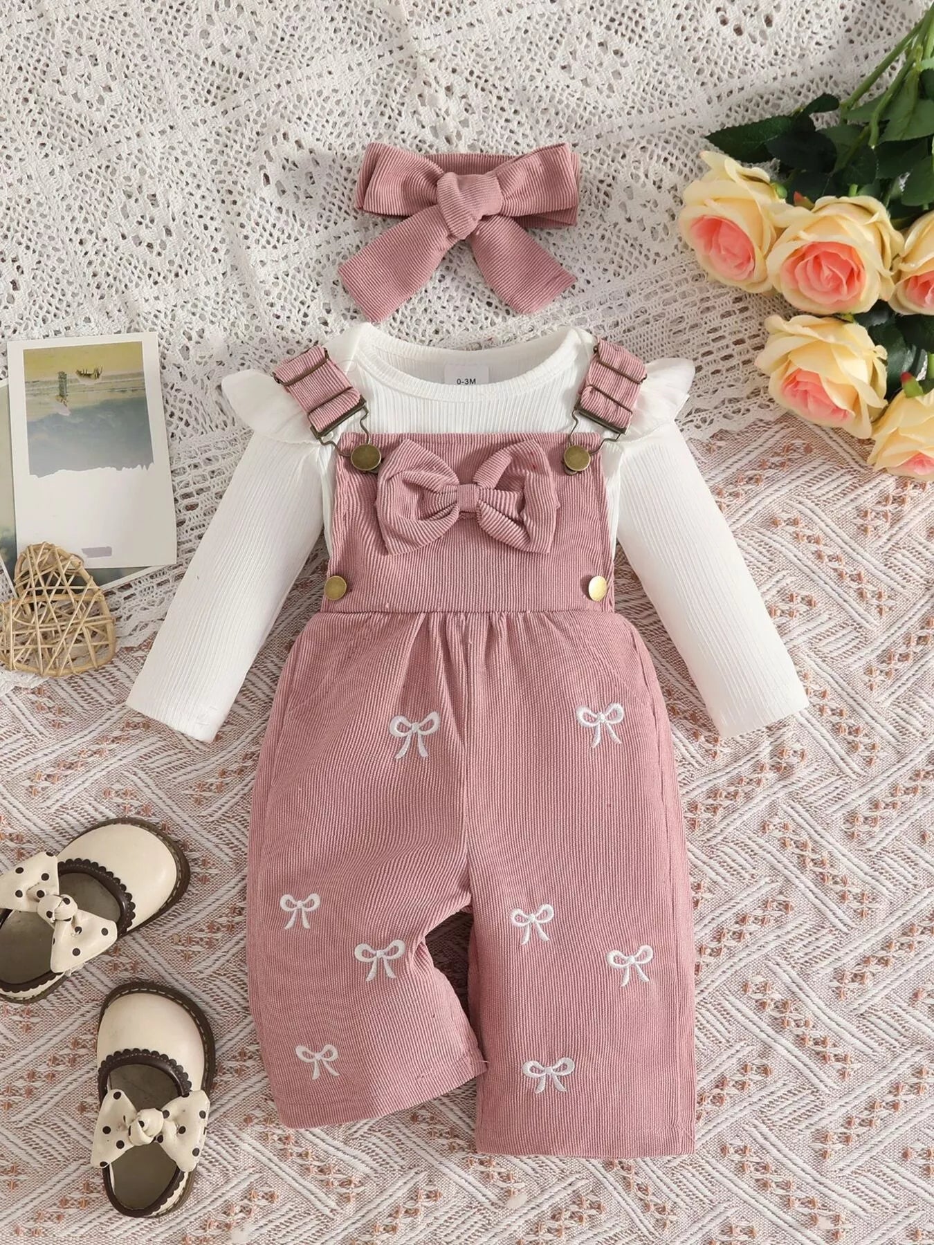 Baby girl three-piece outfit featuring a cream ribbed long-sleeve bodysuit with ruffled shoulders, pink bow-embroidered overalls and matching pink bow headband.