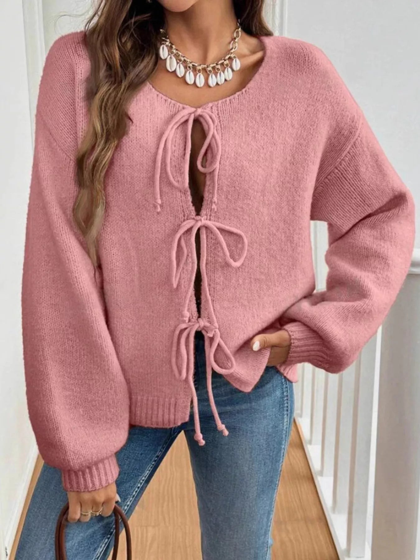 Woman wearing a pink tie-front long sleeve knit cardigan with three bow ties paired with blue jeans.