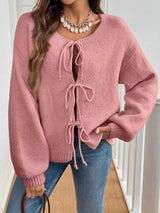 Woman wearing a pink tie-front long sleeve knit cardigan with three bow ties paired with blue jeans.