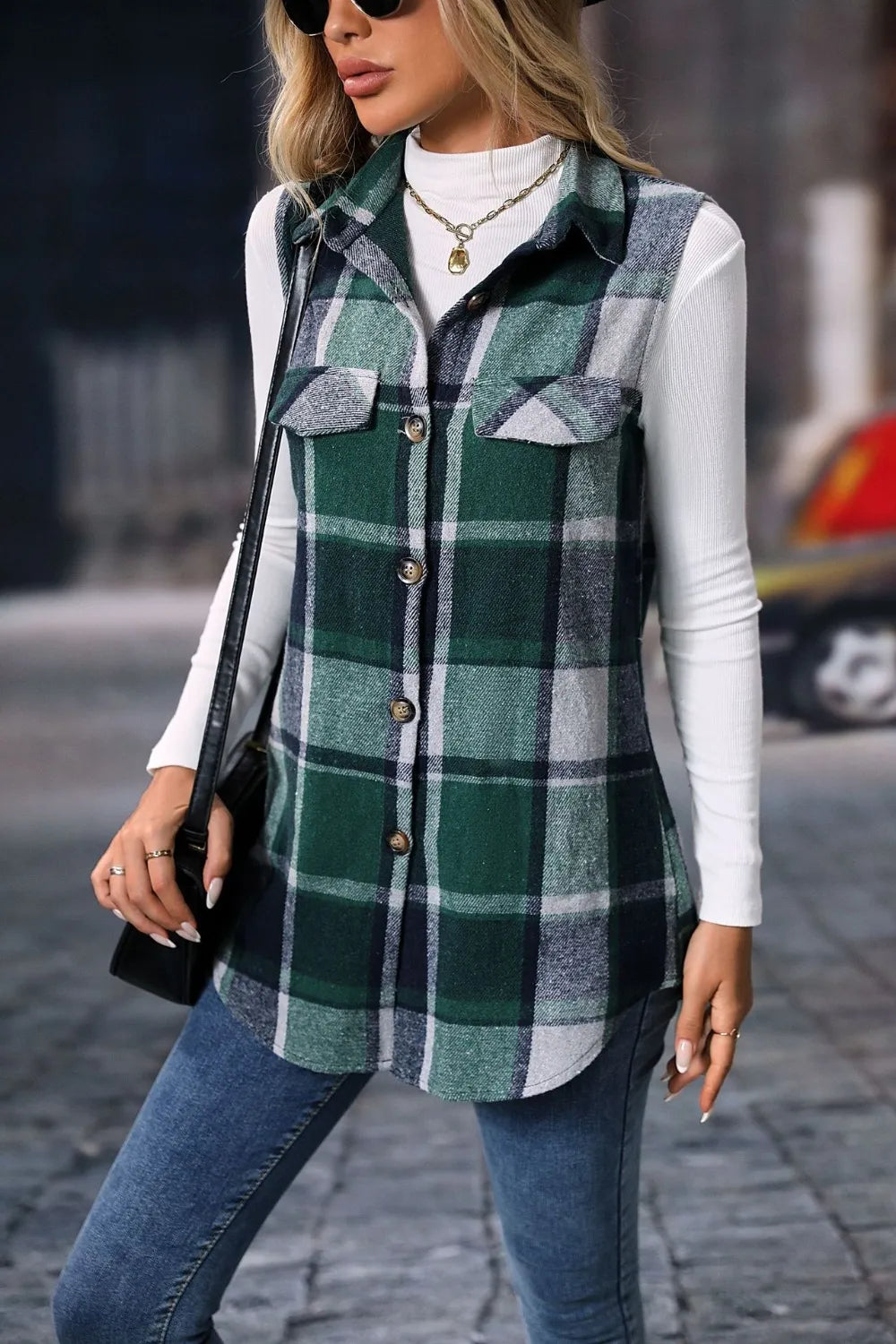 Woman wearing a dark green plaid button-up vest coat layered over a white turtleneck with blue jeans and gold jewelry for a cozy fall outfit.