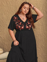 Woman wearing a black dress with red floral embroidery against a textured beige wall.