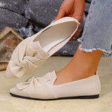 Beige loafers with a bow detail worn by a person against a stone wall background