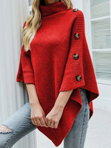 Women's red knit turtleneck poncho pullover with decorative buttons along the sleeves and pointed asymmetrical hem, styled with light-wash jeans.