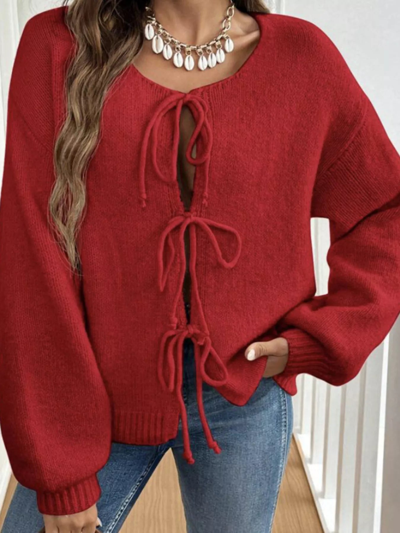 Woman wearing a red tie-front long sleeve knit cardigan with three bow ties paired with blue jeans.